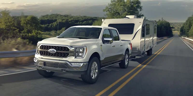 brand new 2023 Ford F-150 towing a smaller, about 22ft, travel trailer up a slightly foresty mountain side highway.