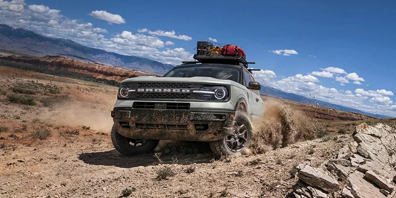 Photo of a brand new pearl white 2023 Ford Bronco Sport with camping gear hitched to the top of it. The Bronco is having a blast kicking up off road desert dirt.
